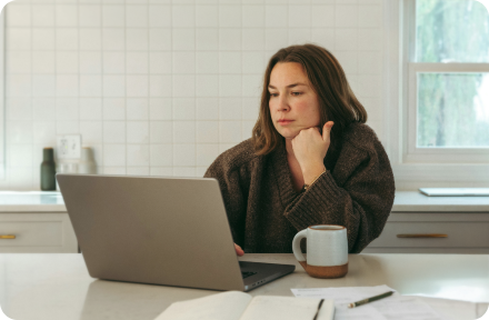 Person sitting in a kitchen, drinking coffee, looking at a laptop with a serious face expression.