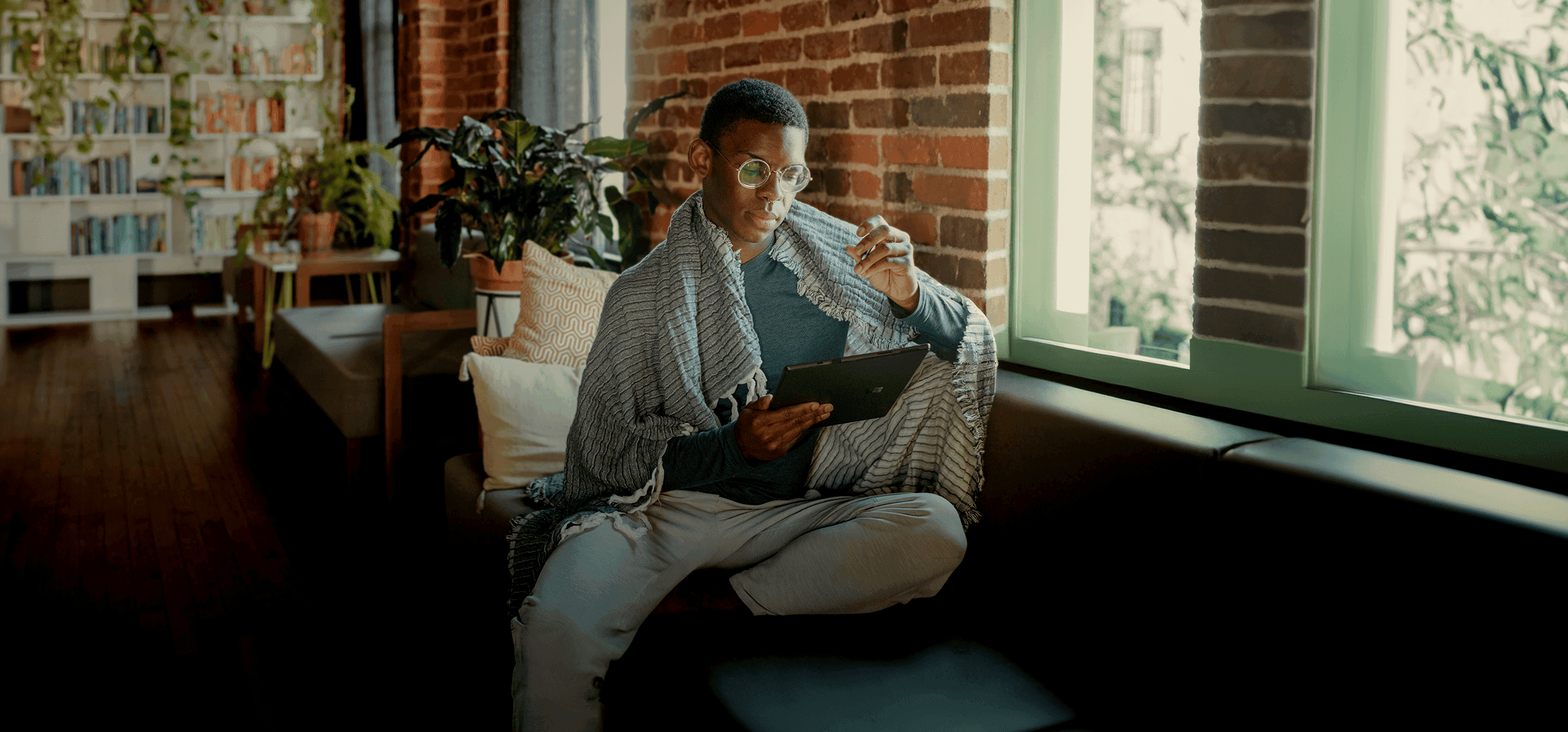 A man with glasses sitting cross-legged on a sofa, wrapped in a blanket and reading from a tablet in a light-filled living room.