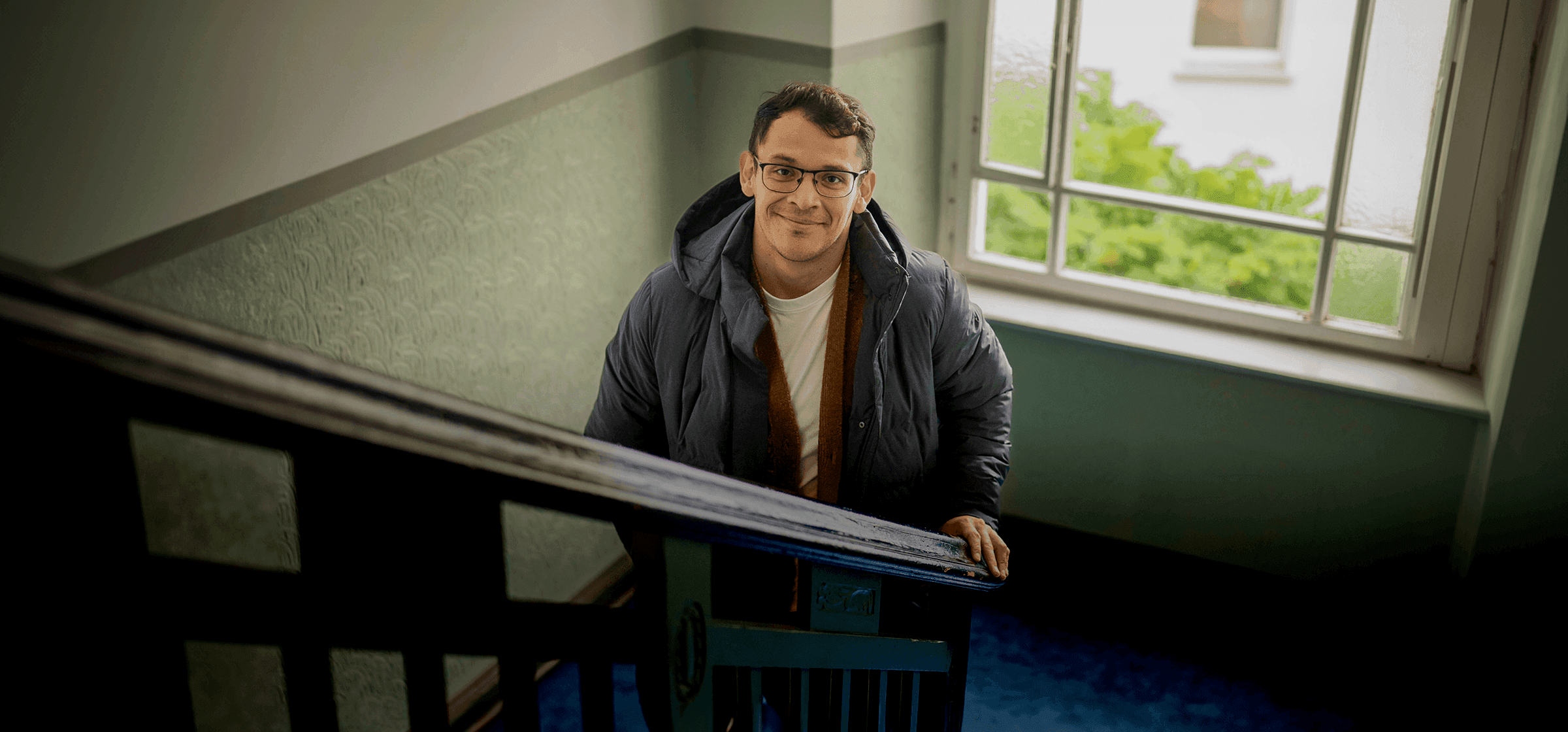 A young man with glasses smiling up at the camera from a stairwell in an apartment building.