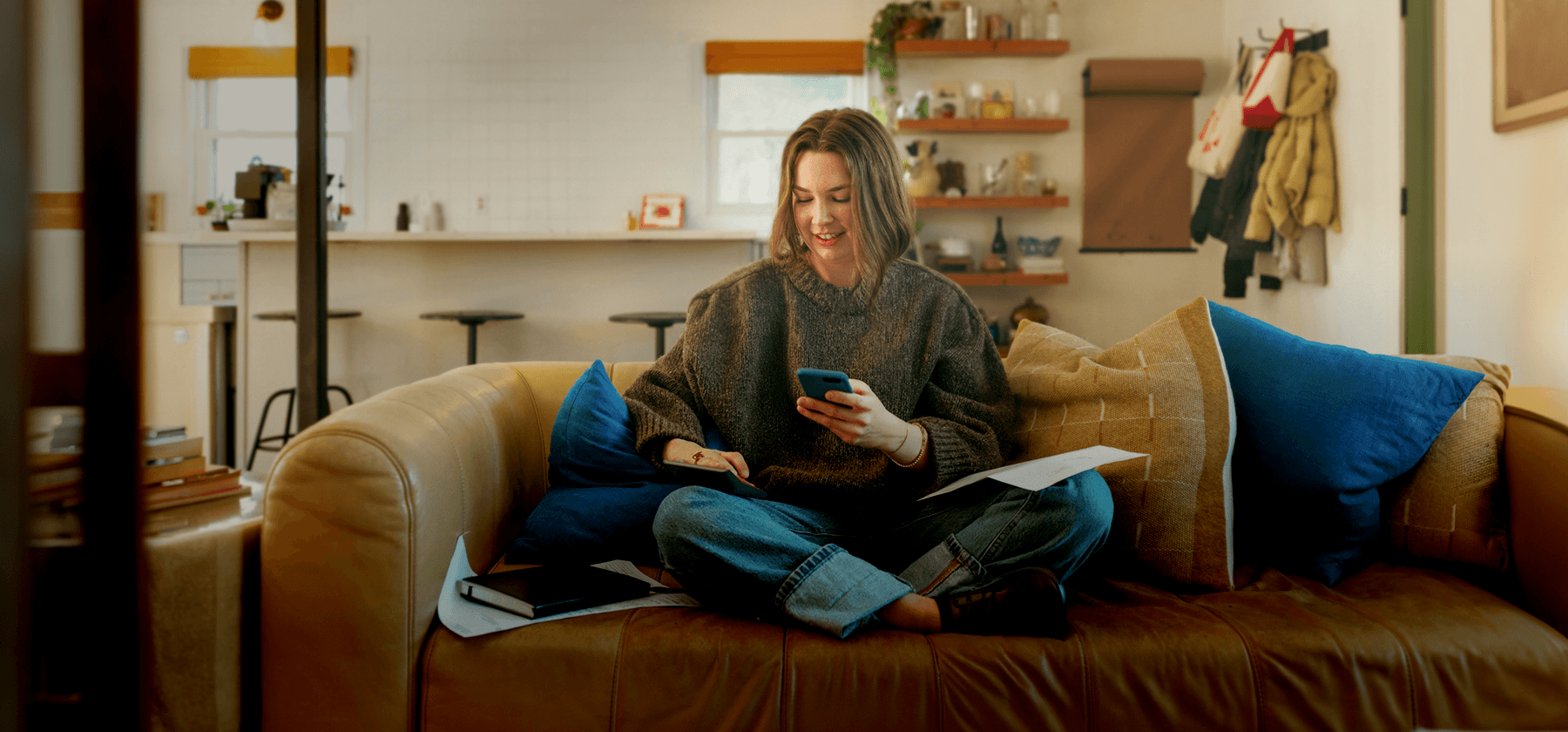 A woman in a chunky knit sweater sitting cross-legged on a leather sofa, smiling at her smartphone with papers and a notebook beside her in Portugal.