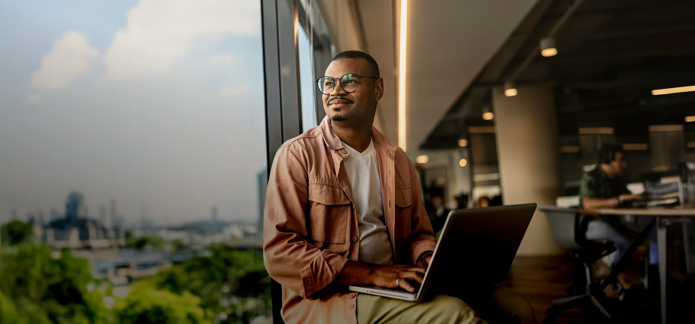A man with glasses sitting with a laptop by a large office window overlooking a city skyline in France