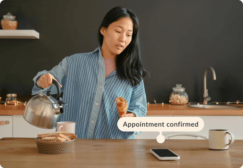 A woman making her breakfast in the kitchen looking at her phone. The tag reads "Appointment confirmed"