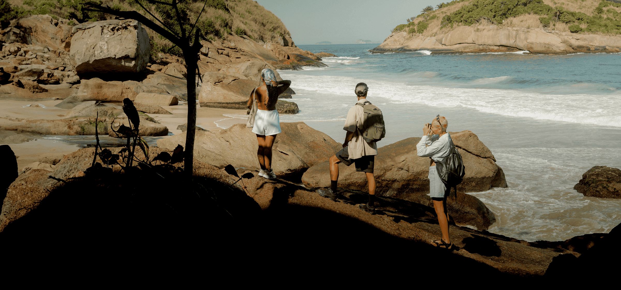 Three travellers with backpacks standing on coastal rocks overlooking a scenic bay with crashing waves in France.